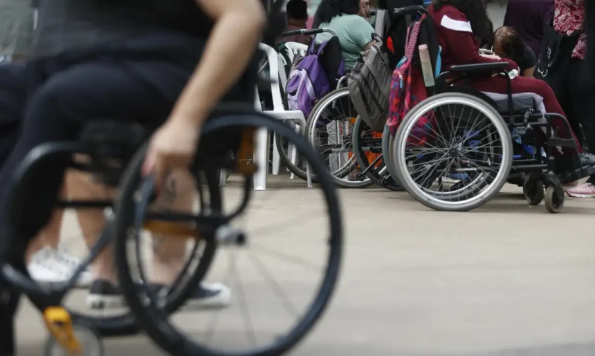 Grupo de pessoas com deficiência usando cadeiras de rodas reunidas em um espaço aberto. Algumas cadeiras possuem mochilas penduradas, sugerindo um ambiente de encontro ou evento. No primeiro plano, uma pessoa em cadeira de rodas manual aparece desfocada.