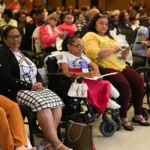 Diversas pessoas, incluindo pessoas com deficiência, participam atentas de um evento ou conferência. Algumas estão em cadeiras de rodas e outras seguram cadernos ou celulares. O ambiente é fechado, bem iluminado e com muitas cadeiras ocupadas. Mercado de Trabalho PCD