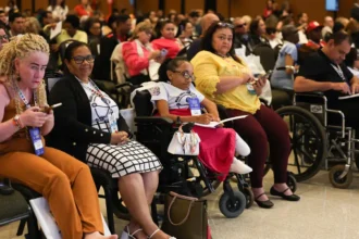 Diversas pessoas, incluindo pessoas com deficiência, participam atentas de um evento ou conferência. Algumas estão em cadeiras de rodas e outras seguram cadernos ou celulares. O ambiente é fechado, bem iluminado e com muitas cadeiras ocupadas. Mercado de Trabalho PCD