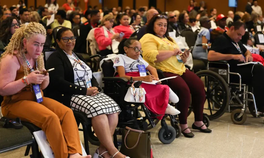 Diversas pessoas, incluindo pessoas com deficiência, participam atentas de um evento ou conferência. Algumas estão em cadeiras de rodas e outras seguram cadernos ou celulares. O ambiente é fechado, bem iluminado e com muitas cadeiras ocupadas. Mercado de Trabalho PCD