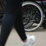 Imagem mostra o foco em rodas de cadeiras de rodas em uma calçada, com destaque para uma pessoa sentada e outra passando em frente, de calça preta e tênis branco.