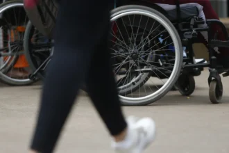 Imagem mostra o foco em rodas de cadeiras de rodas em uma calçada, com destaque para uma pessoa sentada e outra passando em frente, de calça preta e tênis branco.