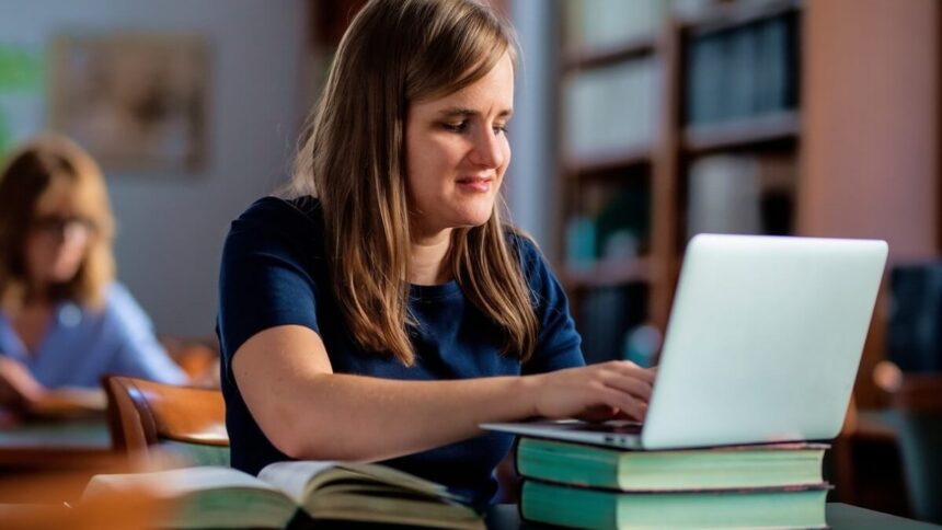 Mulher deficiente visual usando um notebook em uma biblioteca, com livros sobre a mesa. Ela está concentrada no trabalho, e ao fundo há outra pessoa estudando.
