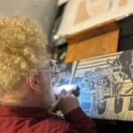 artista plástico Daniel Freita, com cabelos loiros encaracolados e óculos trabalha entalhando uma matriz de xilogravura.