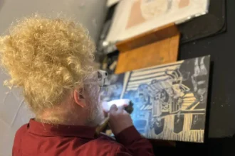 artista plástico Daniel Freita, com cabelos loiros encaracolados e óculos trabalha entalhando uma matriz de xilogravura.