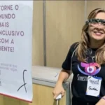 Na imagem, vemos uma mulher sorridente usando um óculos sensorial inovador, projetado para auxiliar pessoas com deficiência visual. Ela veste uma camiseta preta com o logo da iniciativa "SynesthesiAI" e um crachá com o nome "Aida". Ao lado, há um banner que diz: “TORNE O MUNDO MAIS INCLUSIVO COM A GENTE! Conheça o dispositivo sensorial que auxilia a mobilidade de pessoas cegas.”