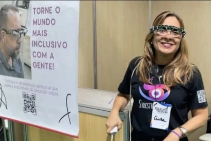 Na imagem, vemos uma mulher sorridente usando um óculos sensorial inovador, projetado para auxiliar pessoas com deficiência visual. Ela veste uma camiseta preta com o logo da iniciativa "SynesthesiAI" e um crachá com o nome "Aida". Ao lado, há um banner que diz: “TORNE O MUNDO MAIS INCLUSIVO COM A GENTE! Conheça o dispositivo sensorial que auxilia a mobilidade de pessoas cegas.”