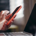 A imagem mostra uma pessoa segurando um smartphone com capa laranja, enquanto digita ou toca na tela, próxima a um notebook aberto. A pessoa usa um relógio inteligente com pulseira azul no pulso esquerdo.