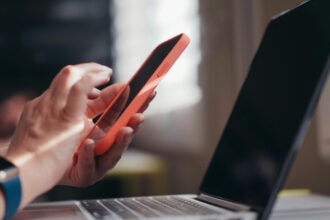 A imagem mostra uma pessoa segurando um smartphone com capa laranja, enquanto digita ou toca na tela, próxima a um notebook aberto. A pessoa usa um relógio inteligente com pulseira azul no pulso esquerdo.