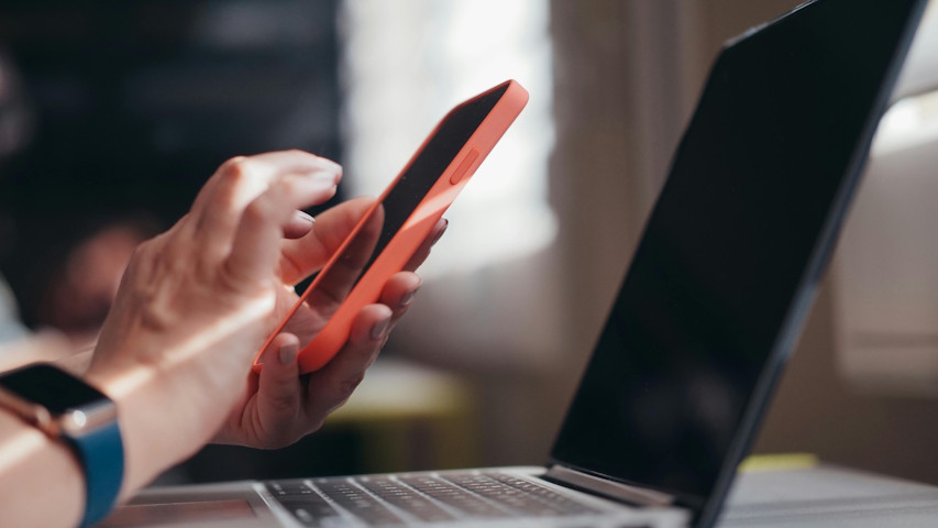 A imagem mostra uma pessoa segurando um smartphone com capa laranja, enquanto digita ou toca na tela, próxima a um notebook aberto. A pessoa usa um relógio inteligente com pulseira azul no pulso esquerdo.