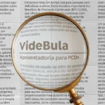 Lupa em destaque ampliando o texto 'VideBula – Aposentadoria para PCDs' sobre uma página de jornal com várias colunas de palavras impressas.