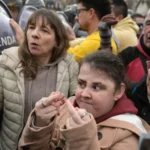 Grupo de manifestantes, incluindo pessoas com deficiência, participa de protesto diante de policiais da Gendarmeria. Uma mulher à frente faz gestos obscenos com os dedos das mãos.