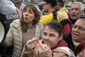 Grupo de manifestantes, incluindo pessoas com deficiência, participa de protesto diante de policiais da Gendarmeria. Uma mulher à frente faz gestos obscenos com os dedos das mãos.
