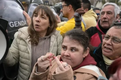Grupo de manifestantes, incluindo pessoas com deficiência, participa de protesto diante de policiais da Gendarmeria. Uma mulher à frente faz gestos obscenos com os dedos das mãos.