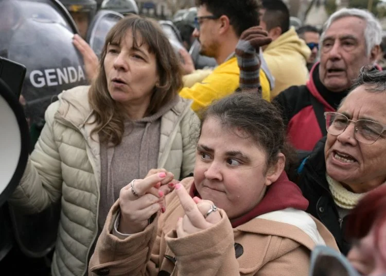 Grupo de manifestantes, incluindo pessoas com deficiência, participa de protesto diante de policiais da Gendarmeria. Uma mulher à frente faz gestos obscenos com os dedos das mãos.