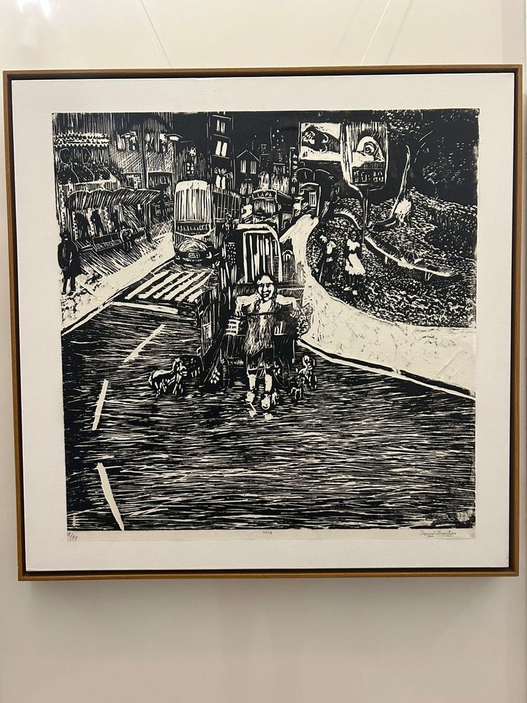 Paula Laboissière/Agência Brasil Gravura em preto e branco mostrando uma cena urbana movimentada. No centro, uma figura humana caminha em meio ao trânsito, ladeada por vários cachorros. Ao redor, vê-se carros, prédios, pessoas nas calçadas e detalhes de um bairro popular. No canto superior direito há um grande outdoor com o rosto de uma mulher. A atmosfera da obra mistura caos urbano e cotidiano.