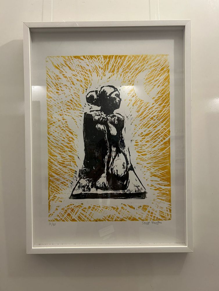 Paula Laboissière/Agência Brasil Gravura emoldurada, mostrando a figura de uma pessoa sentada de perfil, com traços estilizados em preto, sobre um fundo amarelo com linhas radiantes que lembram energia ou luz. No canto superior direito há uma forma de lua crescente. A obra transmite uma sensação de introspecção e força, misturada com intensidade luminosa