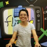 A imagem mostra uma pessoa sorridente, usando óculos redondos e camiseta branca listrada em preto, posicionada em frente a um painel colorido. O painel contém a ilustração de uma televisão antiga com a palavra “flipô” escrita na tela, junto de desenhos variados como um filme cinematográfico, livro com asas, estrelas e figuras geométricas.