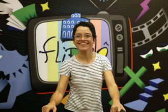 A imagem mostra uma pessoa sorridente, usando óculos redondos e camiseta branca listrada em preto, posicionada em frente a um painel colorido. O painel contém a ilustração de uma televisão antiga com a palavra “flipô” escrita na tela, junto de desenhos variados como um filme cinematográfico, livro com asas, estrelas e figuras geométricas.