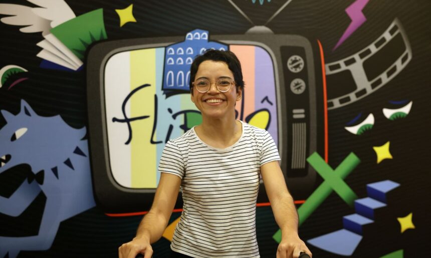 A imagem mostra uma pessoa sorridente, usando óculos redondos e camiseta branca listrada em preto, posicionada em frente a um painel colorido. O painel contém a ilustração de uma televisão antiga com a palavra “flipô” escrita na tela, junto de desenhos variados como um filme cinematográfico, livro com asas, estrelas e figuras geométricas.