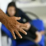 Mão de um homem em primeiro plano, com uma mulher desfocada ao fundo sentada em uma cadeira azul.
