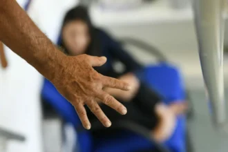 Mão de um homem em primeiro plano, com uma mulher desfocada ao fundo sentada em uma cadeira azul.
