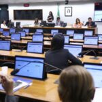 A imagem mostra uma sessão em andamento em uma sala de reuniões oficial, provavelmente de um órgão legislativo ou governamental. Há várias pessoas sentadas diante de mesas equipadas com notebooks ou tablets, enquanto um grupo de autoridades está na mesa da frente. No canto superior direito, há um telão transmitindo a reunião, incluindo intérprete de Libras, o que indica preocupação com acessibilidade para pessoas surdas. No fundo, vê-se a bandeira do Brasil e o retrato oficial de uma autoridade.