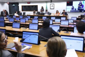 A imagem mostra uma sessão em andamento em uma sala de reuniões oficial, provavelmente de um órgão legislativo ou governamental. Há várias pessoas sentadas diante de mesas equipadas com notebooks ou tablets, enquanto um grupo de autoridades está na mesa da frente. No canto superior direito, há um telão transmitindo a reunião, incluindo intérprete de Libras, o que indica preocupação com acessibilidade para pessoas surdas. No fundo, vê-se a bandeira do Brasil e o retrato oficial de uma autoridade.