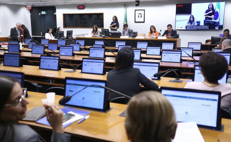 A imagem mostra uma sessão em andamento em uma sala de reuniões oficial, provavelmente de um órgão legislativo ou governamental. Há várias pessoas sentadas diante de mesas equipadas com notebooks ou tablets, enquanto um grupo de autoridades está na mesa da frente. No canto superior direito, há um telão transmitindo a reunião, incluindo intérprete de Libras, o que indica preocupação com acessibilidade para pessoas surdas. No fundo, vê-se a bandeira do Brasil e o retrato oficial de uma autoridade.