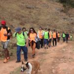Grupo com coletes verdes e laranjas caminha junto por trilha de terra em área montanhosa, acompanhado de um cachorro.