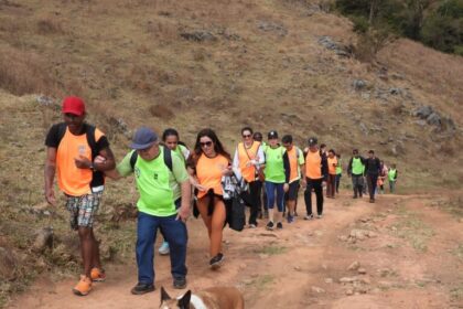 Grupo com coletes verdes e laranjas caminha junto por trilha de terra em área montanhosa, acompanhado de um cachorro.