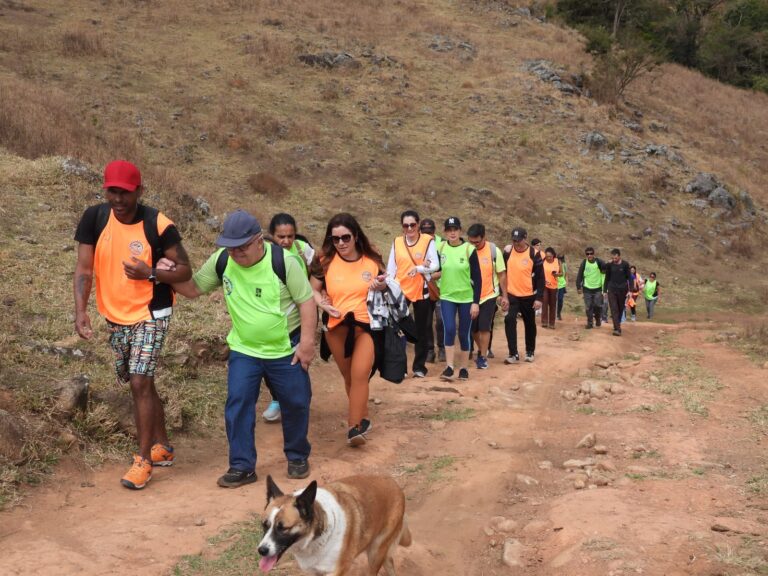 Grupo com coletes verdes e laranjas caminha junto por trilha de terra em área montanhosa, acompanhado de um cachorro.