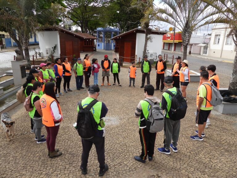 Grupo de pessoas com coletes verdes e laranjas em círculo ao ar livre, participando de trilha inclusiva para a Pedra do coração