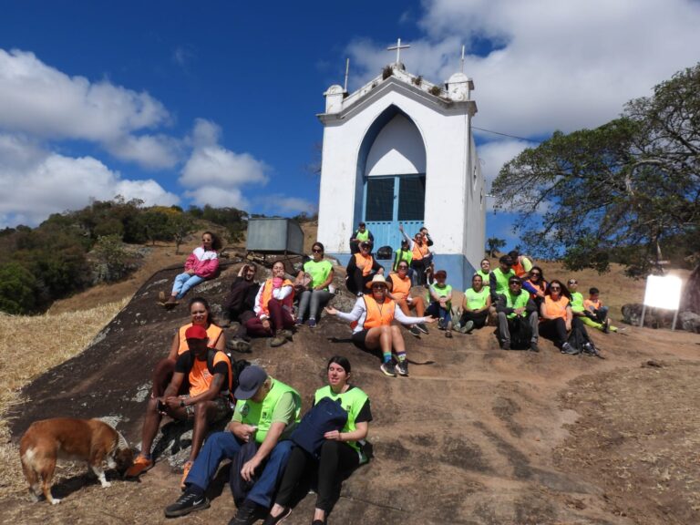 Grupo de pessoas com coletes verdes e laranjas sentado em frente a uma capela durante trilha inclusiva
