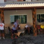 Grupo de pessoas sentadas e em pé em frente a uma construção com varanda e pilares.