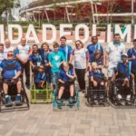 Uma foto de grupo ao ar livre de aproximadamente 20 pessoas, incluindo atletas em cadeiras de rodas motorizadas adaptadas para Power Soccer (Futebol em Cadeira de Rodas) e membros da equipe de apoio ou familiares.