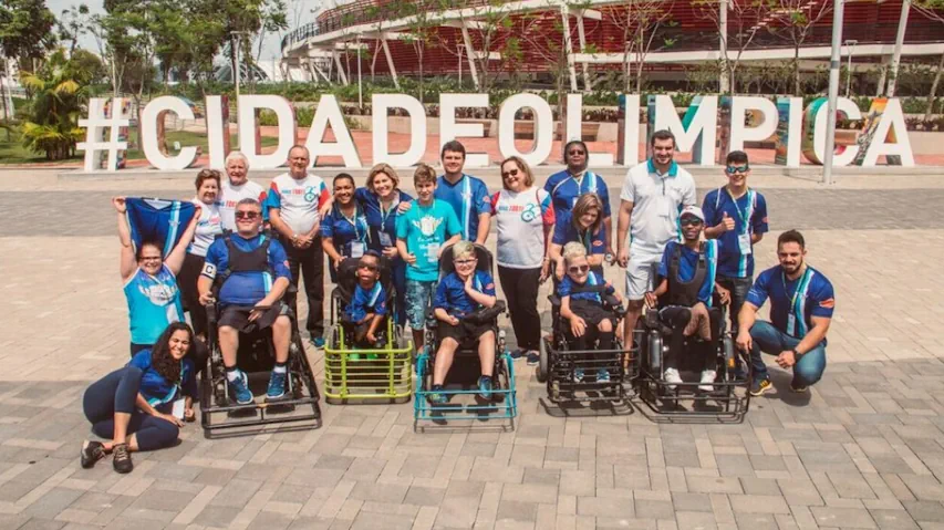 Uma foto de grupo ao ar livre de aproximadamente 20 pessoas, incluindo atletas em cadeiras de rodas motorizadas adaptadas para Power Soccer (Futebol em Cadeira de Rodas) e membros da equipe de apoio ou familiares.