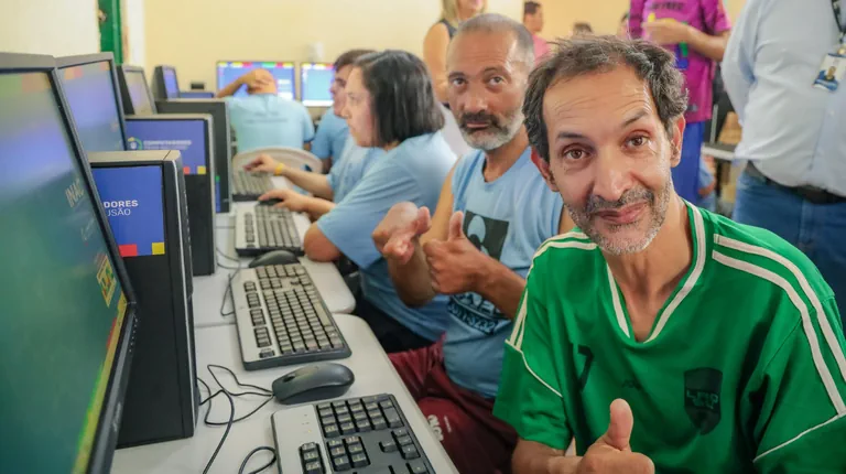 Homens e mulheres com deficiência participam de atividade em um laboratório de informática, utilizando computadores em uma sala equipada. Um dos participantes na frente sorri e faz sinal de positivo para a câmera.