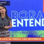Apresentadora da TV Câmara durante o programa ‘Bora Entender’. No rodapé da tela, uma faixa laranja traz a mensagem: ‘Dia da Luta da Pessoa com Deficiência’. Ao fundo, o painel colorido do programa com o título em destaque.
