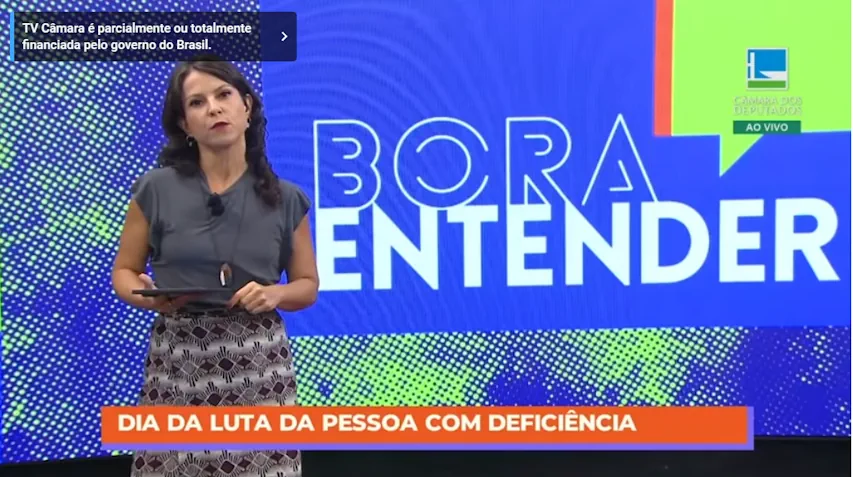 Apresentadora da TV Câmara durante o programa ‘Bora Entender’. No rodapé da tela, uma faixa laranja traz a mensagem: ‘Dia da Luta da Pessoa com Deficiência’. Ao fundo, o painel colorido do programa com o título em destaque.