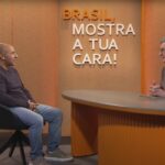 Dois homens participam de uma entrevista em estúdio. Um deles está sentado à esquerda, usando camiseta de manga comprida escura e calça jeans, enquanto o outro está à direita, atrás de uma mesa redonda, vestindo camisa marrom. Ao fundo, na parede laranja, está escrito em letras grandes: ‘Brasil, mostra a tua cara!