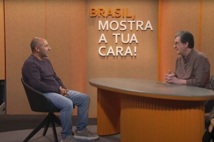 Dois homens participam de uma entrevista em estúdio. Um deles está sentado à esquerda, usando camiseta de manga comprida escura e calça jeans, enquanto o outro está à direita, atrás de uma mesa redonda, vestindo camisa marrom. Ao fundo, na parede laranja, está escrito em letras grandes: ‘Brasil, mostra a tua cara!