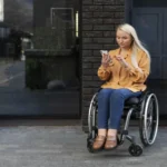 Mulher jovem usuária de cadeira de rodas está sentada em frente a um prédio de tijolos escuros. Ela veste blusa laranja e calça jeans, segura o celular e parece concentrada na tela.