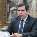 Homem de terno escuro fala ao microfone durante sessão em uma mesa de reuniões. À frente dele há uma placa de identificação com o nome ‘Deputado Ricardo Ayres’. Ao fundo, um quadro grande de estilo clássico.