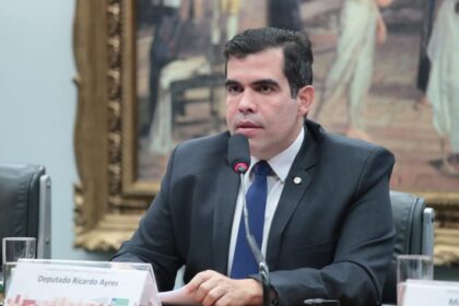 Homem de terno escuro fala ao microfone durante sessão em uma mesa de reuniões. À frente dele há uma placa de identificação com o nome ‘Deputado Ricardo Ayres’. Ao fundo, um quadro grande de estilo clássico.