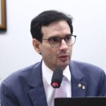 Deputado Léo Prates com pele clara, cabelos pretos curtos e óculos de grau, veste terno azul e camisa branca com gravata lilás. Ele está sentado, falando ao microfone durante uma reunião na Câmara dos Deputados. Ao fundo, parcialmente desfocada, aparece uma mulher de cabelos claros.