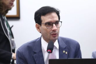 Deputado Léo Prates com pele clara, cabelos pretos curtos e óculos de grau, veste terno azul e camisa branca com gravata lilás. Ele está sentado, falando ao microfone durante uma reunião na Câmara dos Deputados. Ao fundo, parcialmente desfocada, aparece uma mulher de cabelos claros.