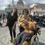 Homem em cadeira de rodas, usando blusa amarela, posa sorridente ao lado de duas mulheres em frente à Praça de São Pedro, no Vaticano. Ao fundo, a Basílica de São Pedro e várias pessoas circulando pelo local.