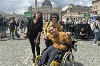 Homem em cadeira de rodas, usando blusa amarela, posa sorridente ao lado de duas mulheres em frente à Praça de São Pedro, no Vaticano. Ao fundo, a Basílica de São Pedro e várias pessoas circulando pelo local.