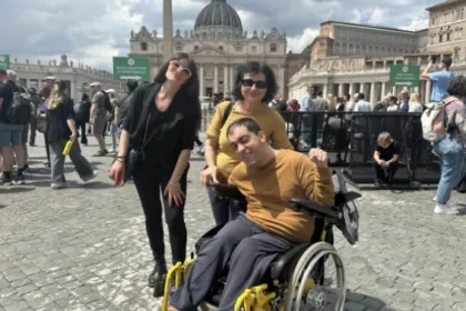 Homem em cadeira de rodas, usando blusa amarela, posa sorridente ao lado de duas mulheres em frente à Praça de São Pedro, no Vaticano. Ao fundo, a Basílica de São Pedro e várias pessoas circulando pelo local.